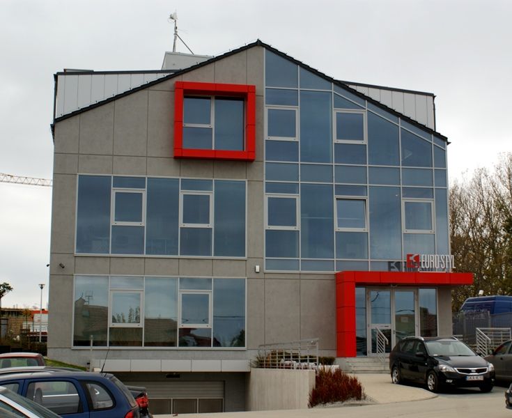 Nowoczesny, trzypiętrowy budynek z szarą fasadą, dużymi szklanymi oknami, czerwoną kwadratową ramą okienną i czerwonym, kanciastym wejściem z logo „EURO STYL”.