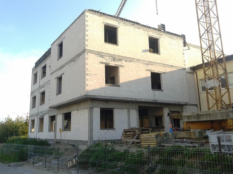 Trzypiętrowy budynek w budowie, zbudowany z jasnych bloków i pustych otworów okiennych, obok dużego dźwigu budowlanego.