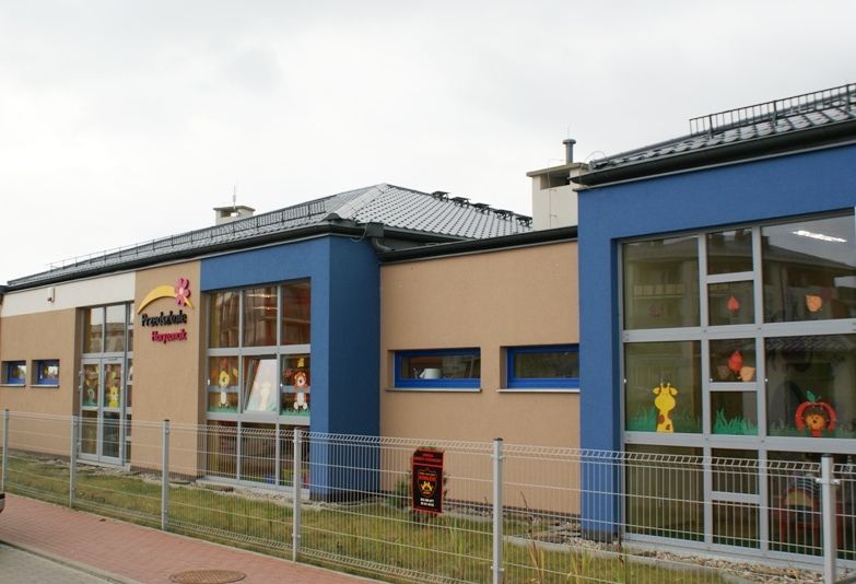 Nowoczesny budynek przedszkola z jasnobrązowymi i niebieskimi ścianami, dużymi oknami ozdobionymi rysunkowymi zwierzętami i płotem z przodu.