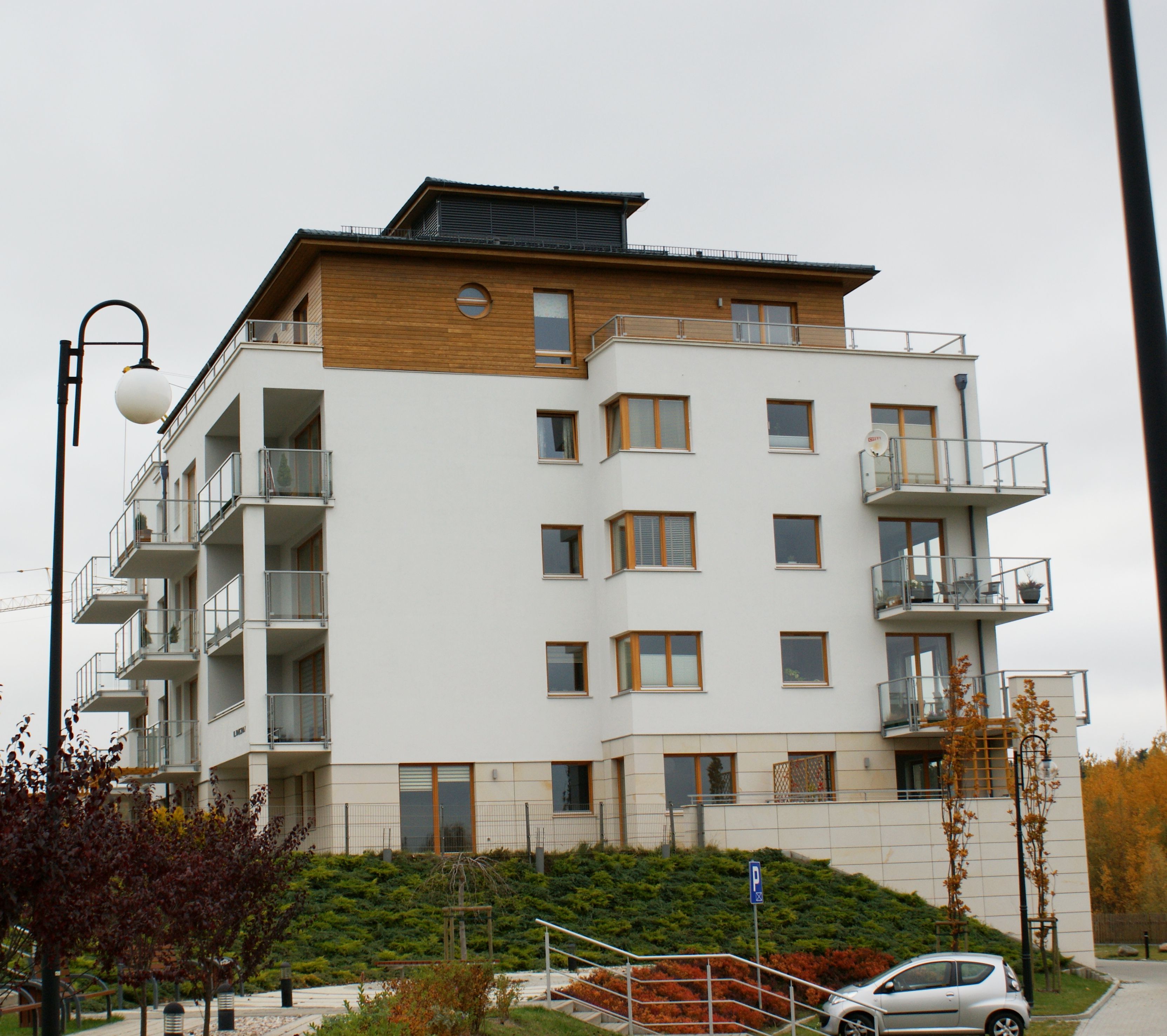 Nowoczesny biały budynek mieszkalny z drewnianymi panelami, szklanymi balkonami i zieloną roślinnością pod zachmurzonym niebem.