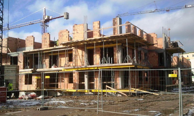 Wielopiętrowy budynek z cegły i betonu w budowie, z rusztowaniem, dźwigiem i tymczasowym ogrodzeniem na błotnistym gruncie.