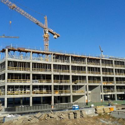 A multi-story concrete building under construction with a large crane lifting a beam against a clear blue sky.