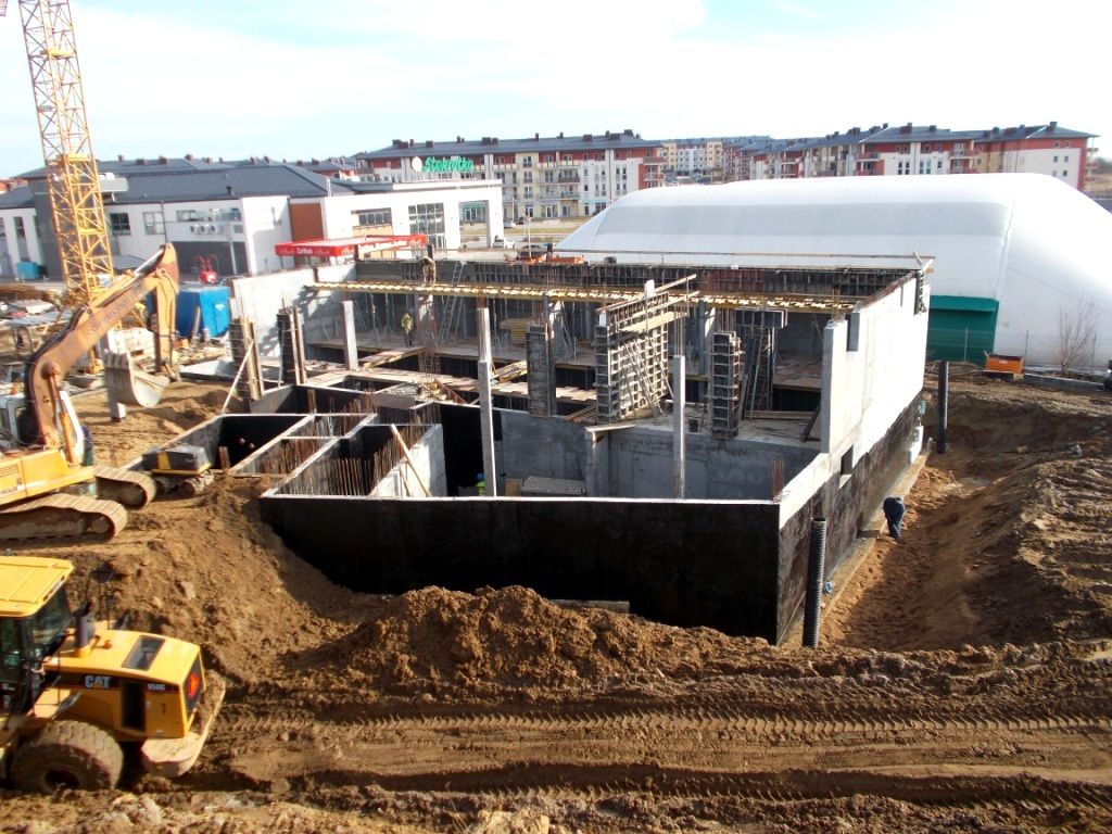 Budynek w budowie z odsłoniętym betonowym fundamentem i ścianami, otoczony ziemią, koparkami i dźwigiem, z budynkami mieszkalnymi i białą kopułą w tle.