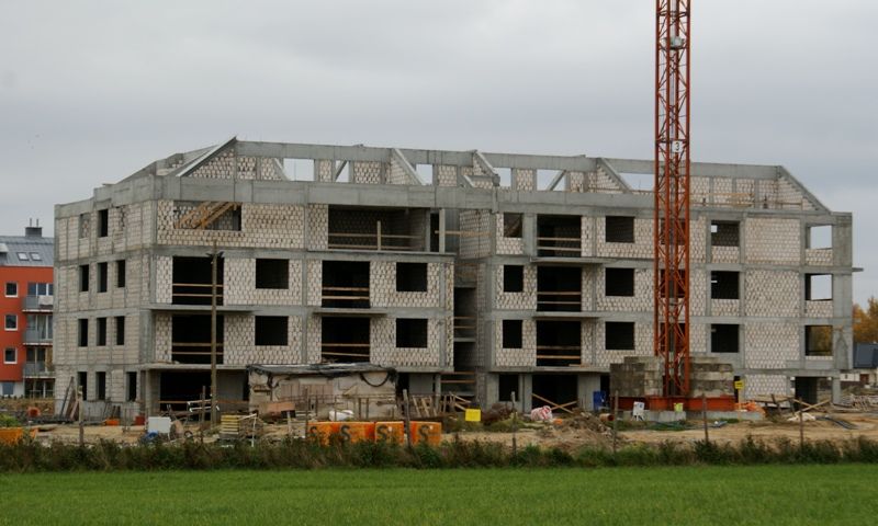 Wielopiętrowy betonowy budynek w budowie z pomarańczowym dźwigiem i zielonym polem.