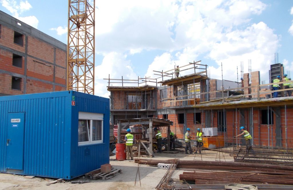 Robotnicy na placu budowy, wokół wznoszonych budynków, niebieskiego przenośnego biura i żółtego dźwigu.