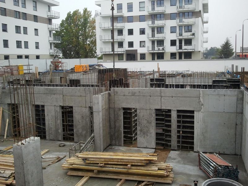 Betonowe ściany fundamentów na czynnym placu budowy. W tle widać nowoczesne, białe budynki mieszkalne.