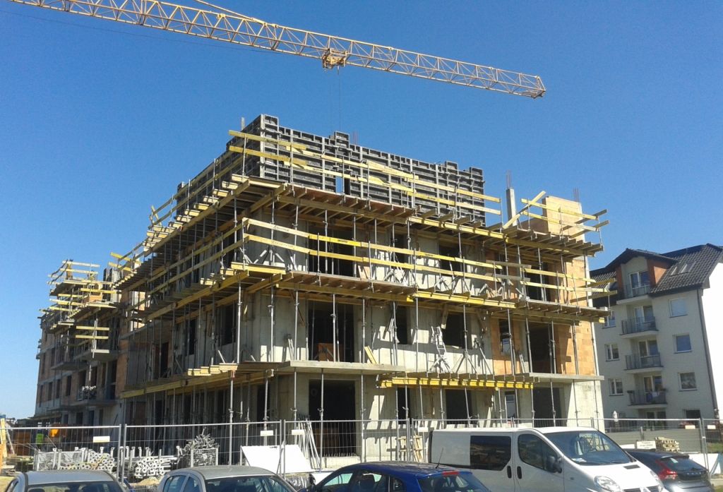 Wielopiętrowy budynek w budowie, z rusztowaniami i dźwigiem pod czystym, błękitnym niebem.
