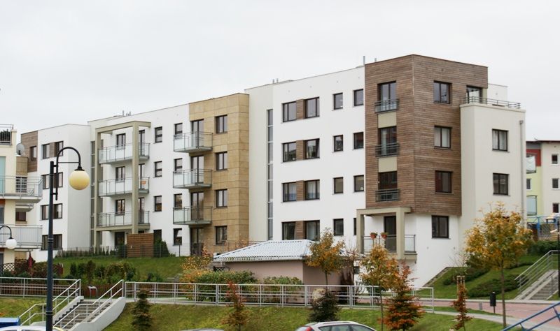 Nowoczesny budynek apartamentowy z białymi i fakturowanymi fasadami, balkonami, na zielonym zboczu z jesiennymi drzewami.