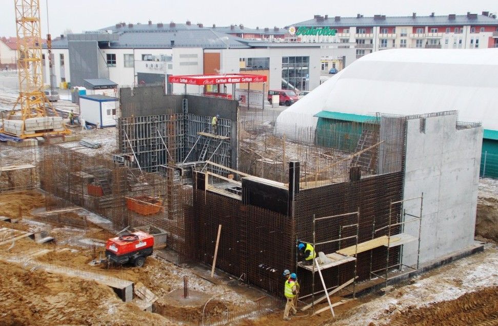 Plac budowy z odsłoniętymi prętami zbrojeniowymi, betonowymi ścianami, pracownikami, dźwigiem i budynkami w tle.