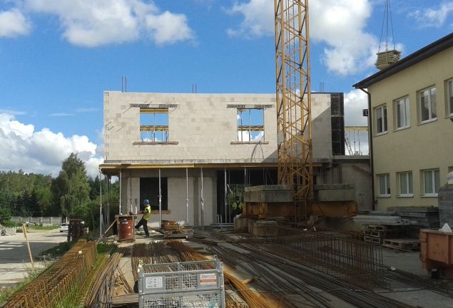 Plac budowy z częściowo wzniesionym budynkiem z bloków, żółtym dźwigiem, robotnikiem i materiałami budowlanymi pod błękitnym niebem.