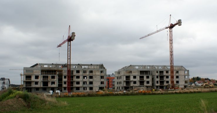 Dwa niedokończone betonowe budynki z dźwigami na polu pod zachmurzonym niebem.