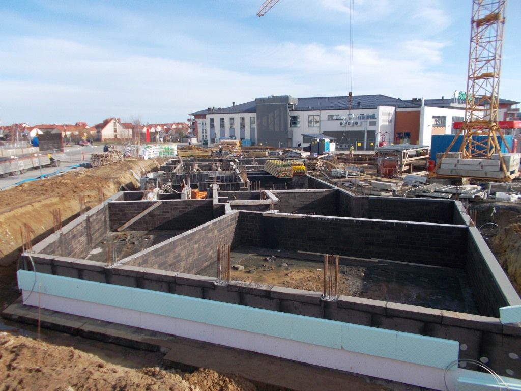 Fundamenty budynku w budowie, na pierwszym planie ściany z bloczków i izolacja, w tle dźwig i inne budynki.