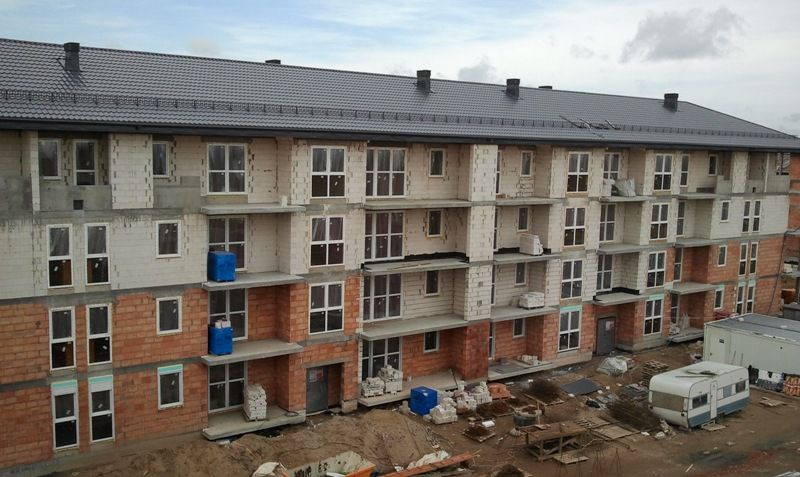 Wielopiętrowy budynek mieszkalny w trakcie budowy z czerwonymi ceglanymi i jasnymi bloczkami ściennymi, ciemnym dachem i placem budowy poniżej.