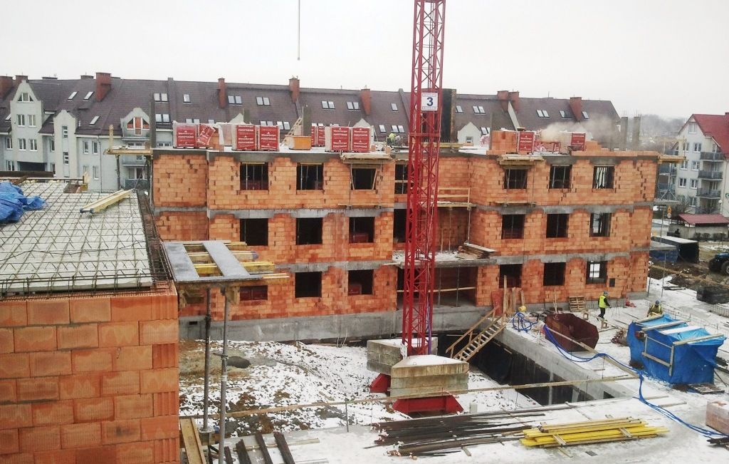 Plac budowy z częściowo wzniesionym budynkiem z cegły, czerwonym dźwigiem i śniegiem na ziemi.