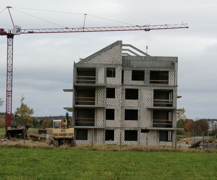 Czteropiętrowy budynek w budowie z czerwonym dźwigiem i żółtą koparką, położony na zielonym polu pod zachmurzonym niebem.