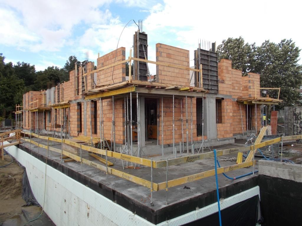 Wielopiętrowy budynek w trakcie budowy, z częściowo zbudowanymi ścianami z cegły, szalunkami betonowymi i rusztowaniem.