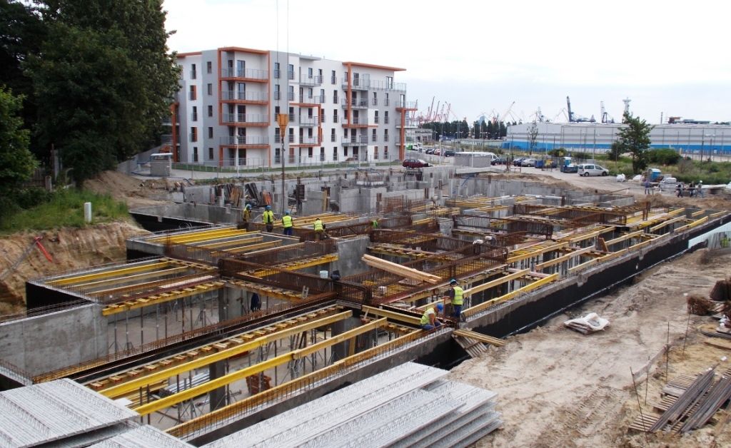 Robotnicy budowlani budujący betonowe fundamenty i niższe piętra nowego budynku, w oddali widać kompleks apartamentów i port.