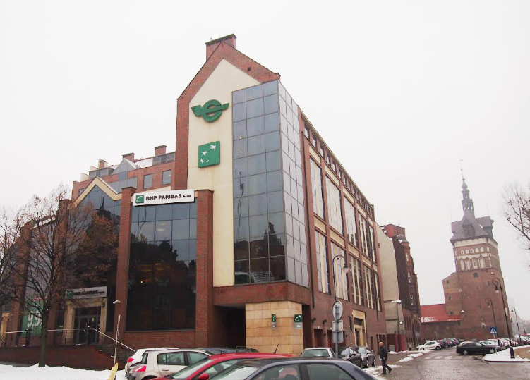 Nowoczesny budynek banku BNP Paribas na zaśnieżonej ulicy ze starą ceglaną wieżą w tle.