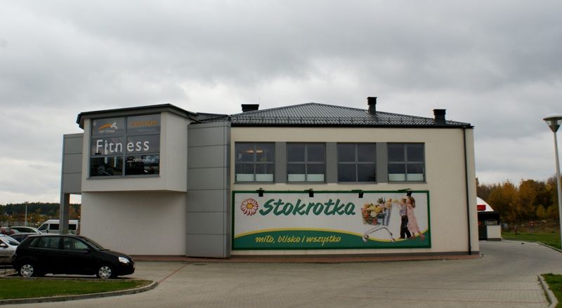 Budynek mieszczący centrum fitness i supermarket Stokrotka z parkingiem pod zachmurzonym niebem.