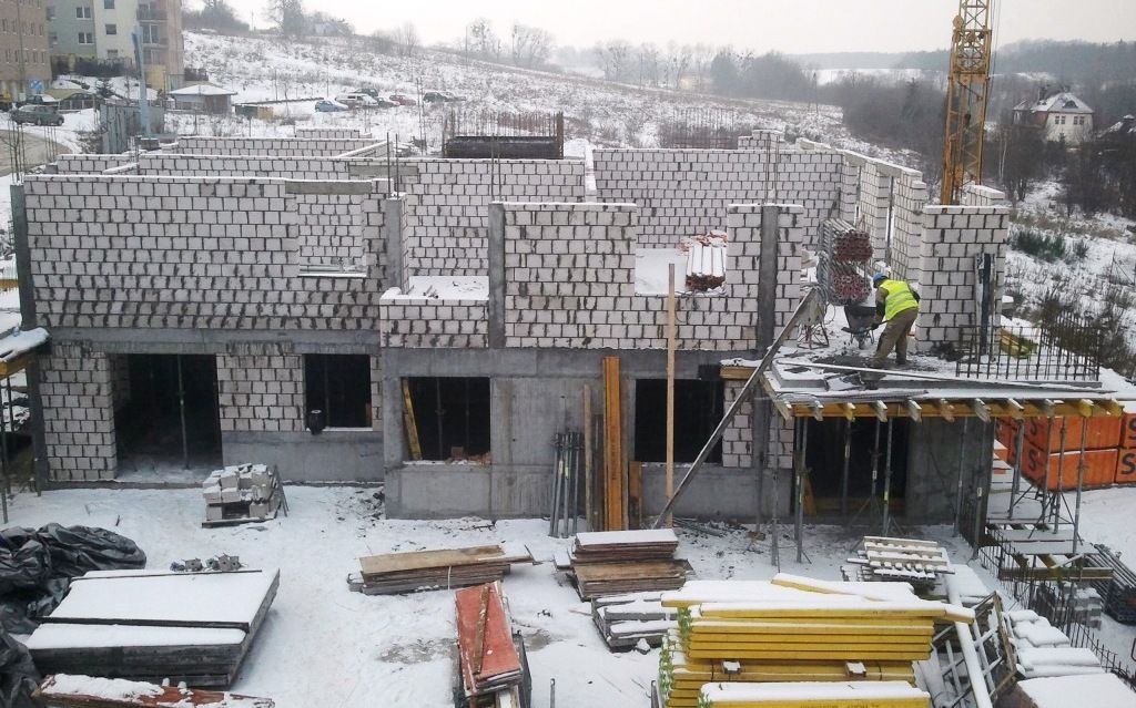 Robotnik budowlany obsługuje dźwig na placu budowy, na którym powstają ściany z bloczków betonowych, w otoczeniu śniegu i materiałów.