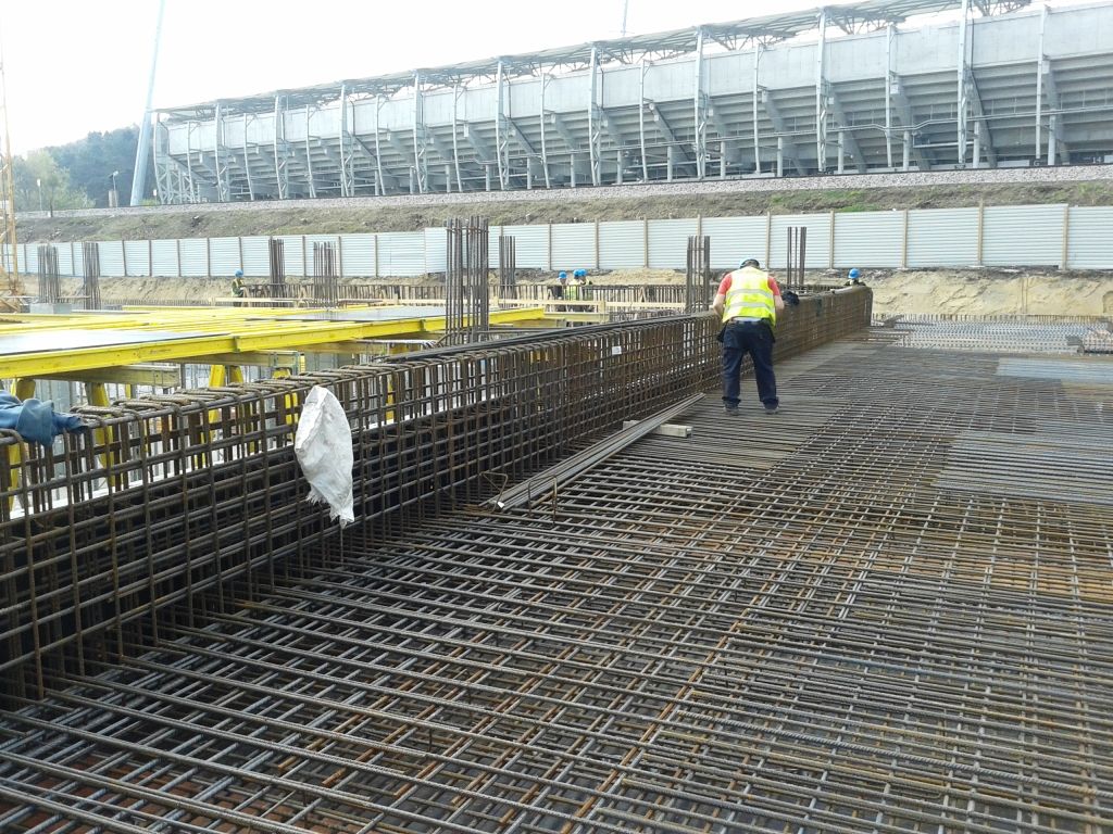 Plac budowy ze stalowymi prętami zbrojeniowymi, pracownikiem i stadionem w tle.