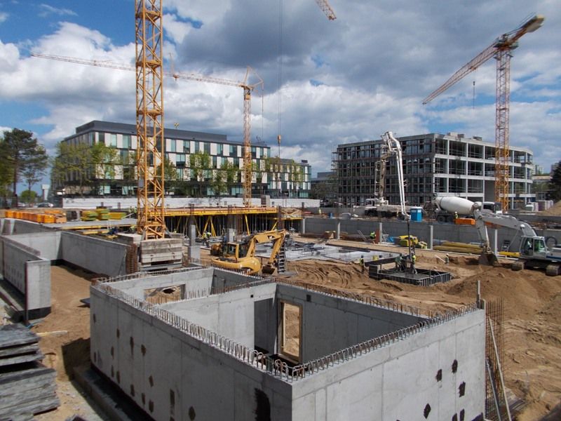 Plac budowy z betonowymi fundamentami, wieloma dźwigami, ciężkim sprzętem i budynkami w trakcie budowy.