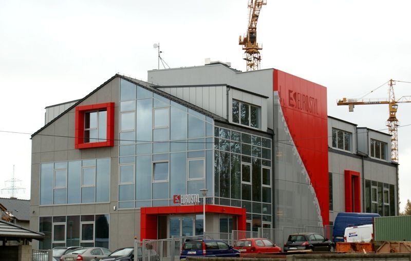 Nowoczesny szaro-czerwony budynek ze szklaną fasadą i napisem „EUROSTYL” na tle dźwigów budowlanych.