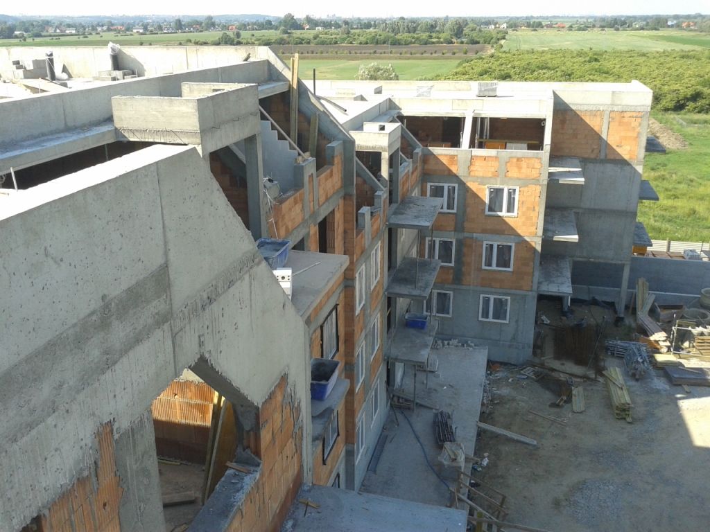 Wielopiętrowy budynek w trakcie budowy z odsłoniętym betonem, ceglanymi ścianami i zamontowanymi oknami, położony na tle zielonych pól.