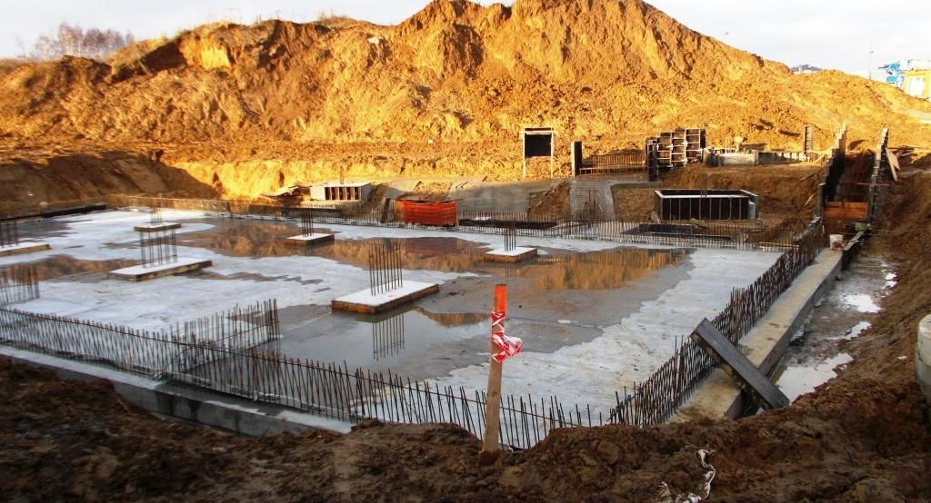 Fundamenty placu budowy ze stojącą wodą, prętami zbrojeniowymi i stopami fundamentowymi, otoczone dużymi kopcami ziemi.