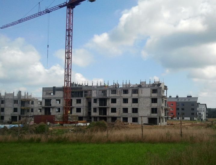 Wielopiętrowe budynki w budowie z dużym dźwigiem, widziane z trawiastego pola.