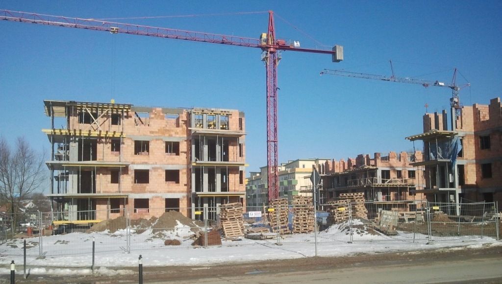 Niedokończone ceglane budynki i czerwone dźwigi wieżowe na zaśnieżonym placu budowy pod czystym, błękitnym niebem.