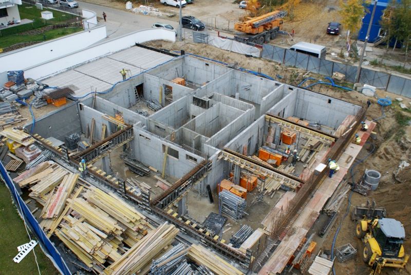 Widok z lotu ptaka na betonowy fundament wielopomieszczeniowego budynku w trakcie budowy, z pracownikami, materiałami budowlanymi i ciężkim sprzętem.