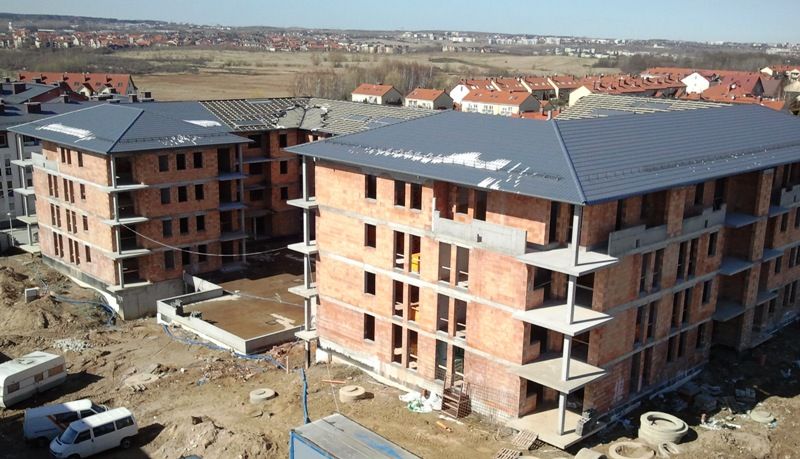 Dwa ceglane budynki mieszkalne z ciemnoszarymi dachami w budowie, położone na ziemnej działce z innymi budynkami mieszkalnymi w tle.