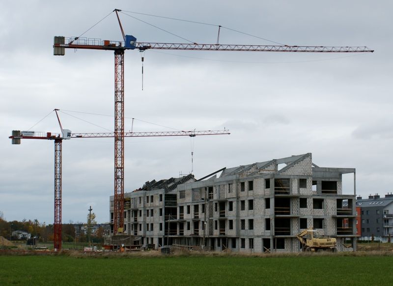 Dwa czerwone dźwigi wieżowe nadzorują budowę wielopiętrowego budynku, w pobliżu stoi koparka, a na pierwszym planie widać zieloną trawę.