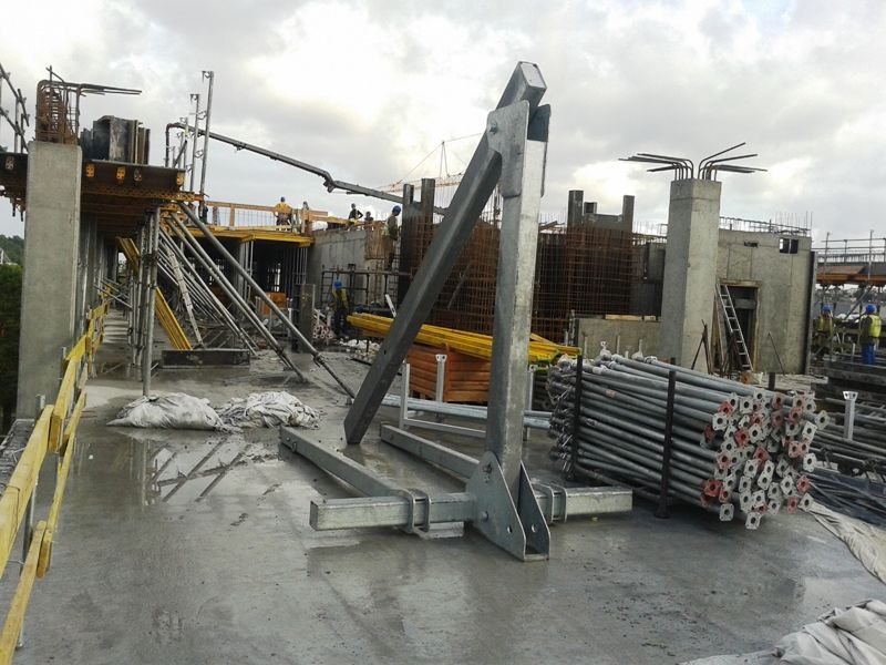 Plac budowy z metalową ramą i ułożonymi w stosy słupami rusztowania na mokrej betonowej podłodze. W tle widoczne są konstrukcje budynków i pracownicy.