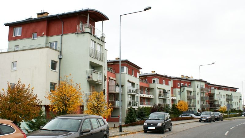 Szereg nowoczesnych budynków mieszkalnych z czerwonymi dachami i balkonami wzdłuż ulicy z samochodami i jesiennymi drzewami.