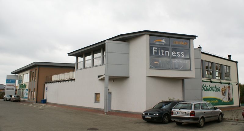 Budynek z centrum fitness i sklepem spożywczym Stokrotka, przed którym, pod zachmurzonym niebem, zaparkowane są samochody.