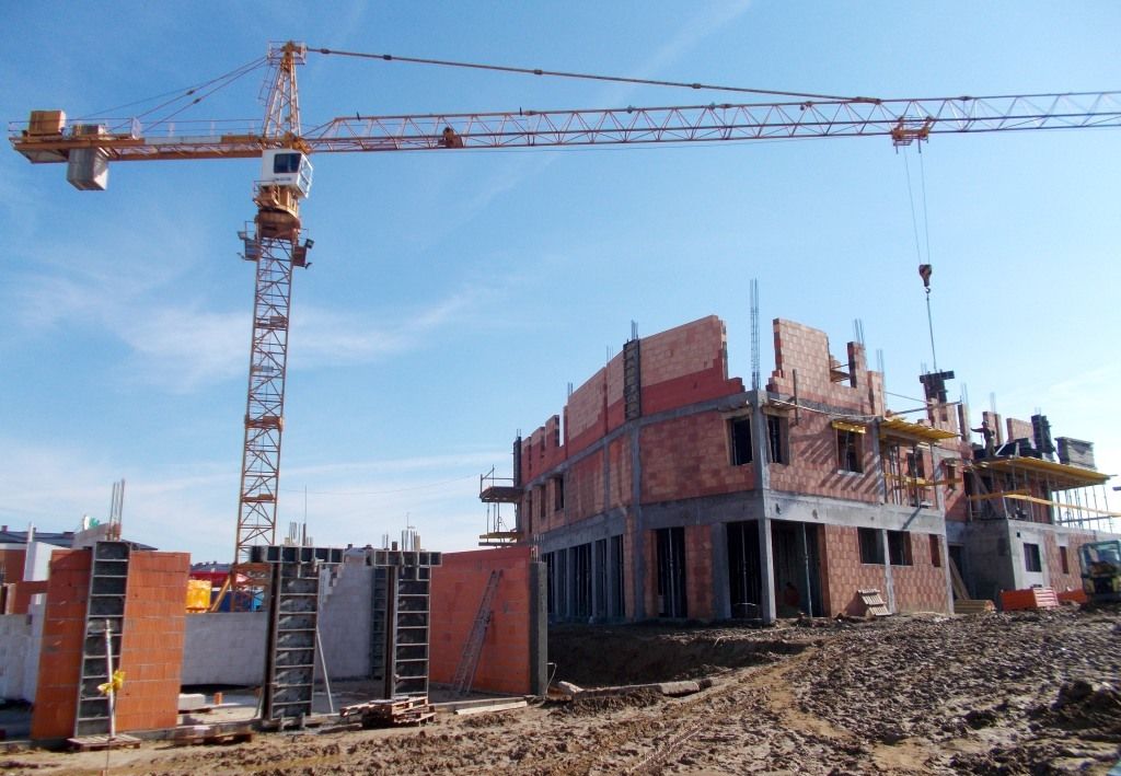 Plac budowy z dźwigiem wieżowym i częściowo wybudowanymi budynkami z cegły.