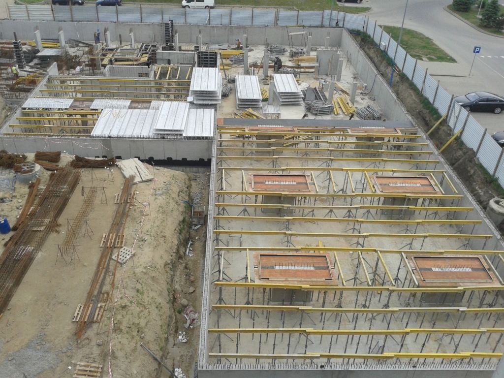 Widok z góry na plac budowy z betonowymi ścianami fundamentowymi, deskowaniem płyty stropowej i materiałami budowlanymi.