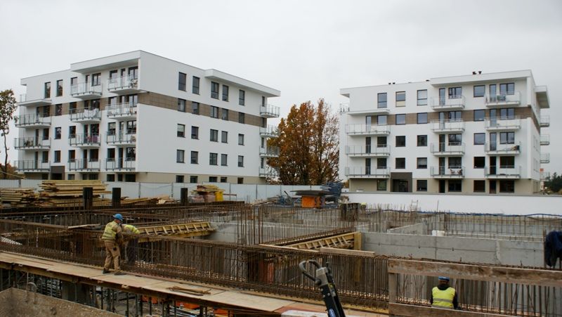 Plac budowy z robotnikami i odsłoniętymi betonowymi fundamentami. W tle dwa nowoczesne białe budynki mieszkalne.