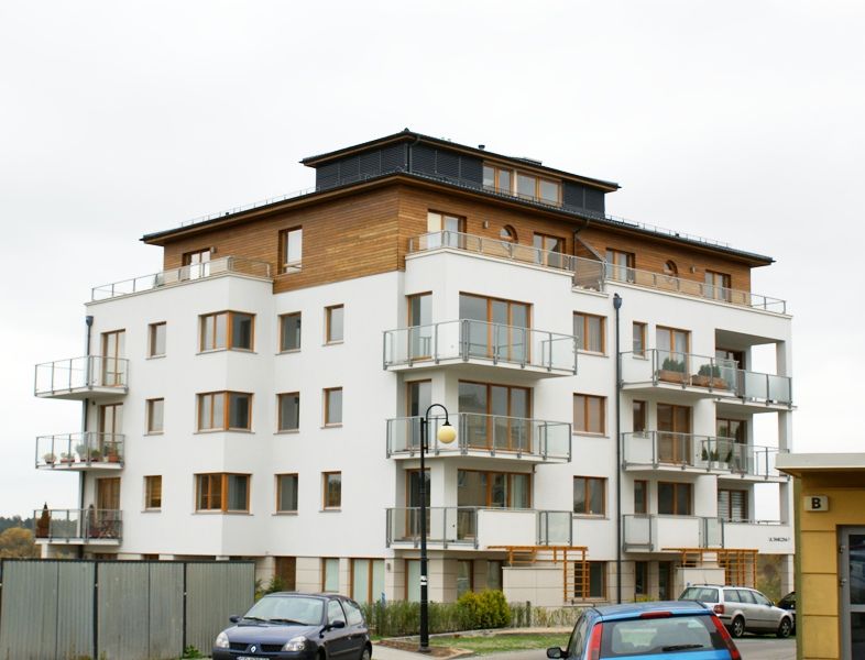 Nowoczesny biały budynek apartamentowy z drewnianym ostatnim piętrem i balkonami z przeszklonymi balustradami.