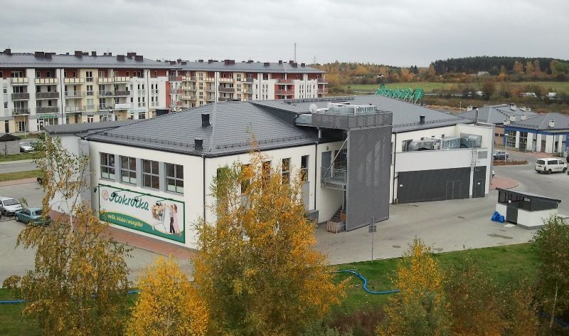 Biały budynek komercyjny ze znakiem „Rokrokka”, otoczony jesiennymi drzewami i budynkami mieszkalnymi, pod zachmurzonym niebem.