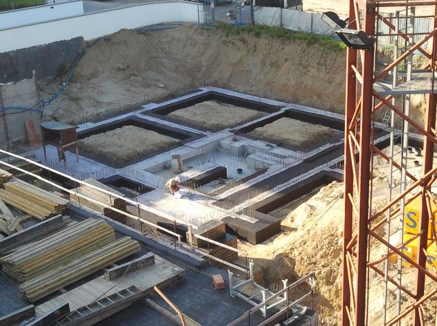 Trwające prace fundamentowe w wykopie na placu budowy. Po prawej stronie widoczny jest dźwig.