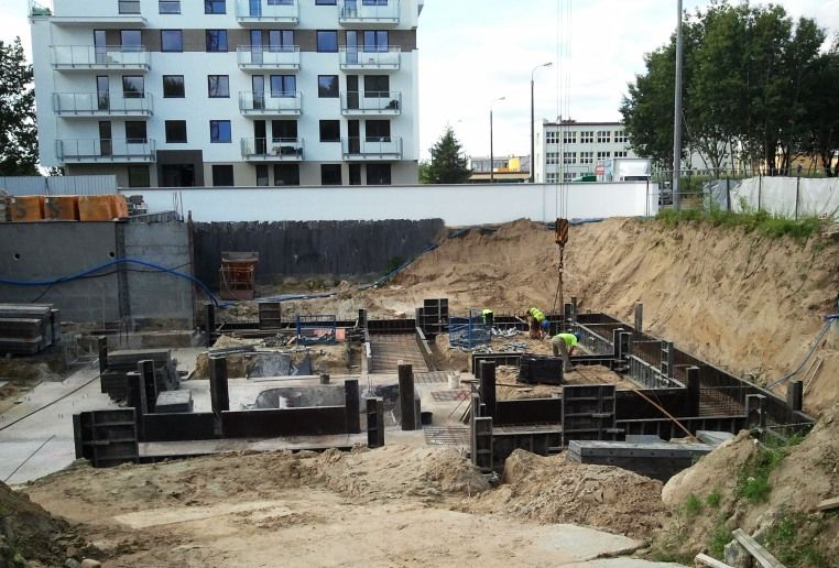 Wykopany dół fundamentowy, na którym robotnicy stawiają szalunki, a w oddali widać gotowy budynek mieszkalny.