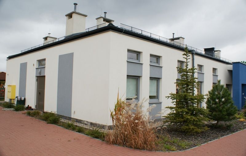 Nowoczesny, parterowy, biały budynek z szarymi akcentami, otoczony zielenią i czerwoną ceglaną ścieżką, pod zachmurzonym niebem.