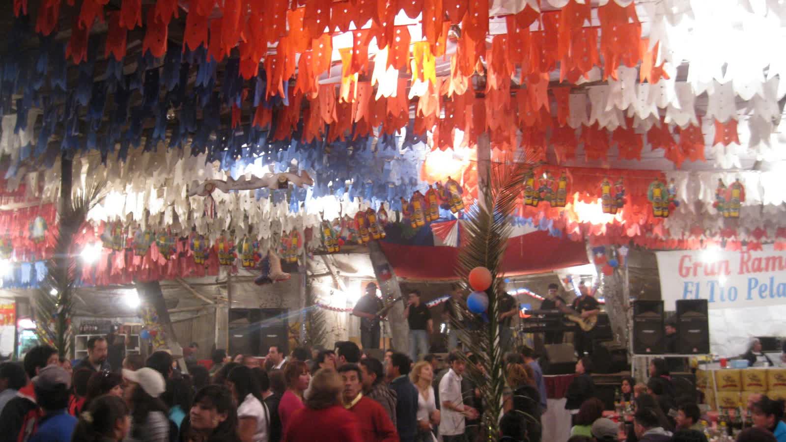 Personas bailando bajo guirnaldas de papel en una fonda de Fiestas Patrias, con una orquesta tocando al fondo