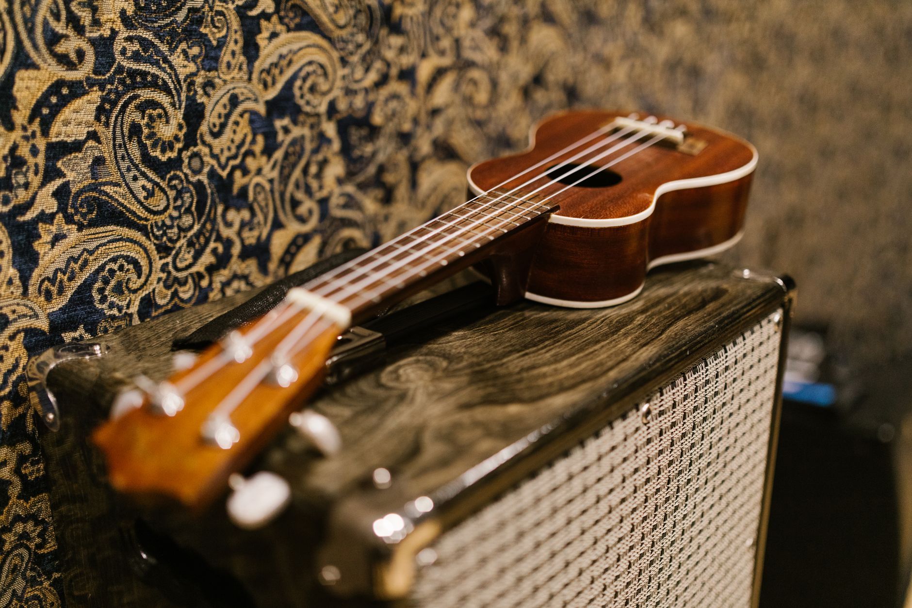 Imagen referencial: Acercamiento de un ukulele sobre un amplificador vintage frente a un fondo de papel tapiz ornamentado.