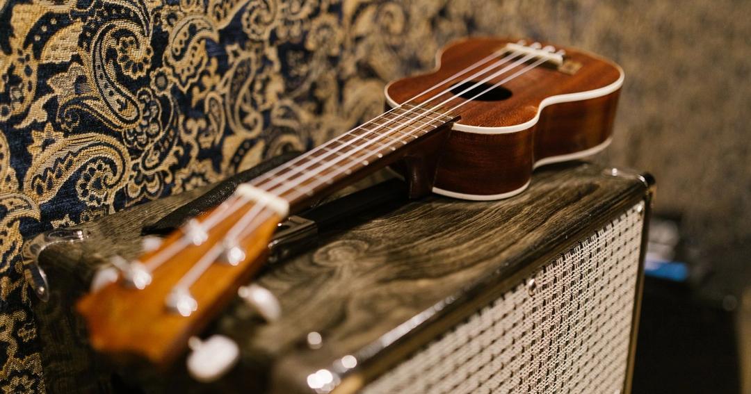 Imagen referencial: Acercamiento de un ukulele sobre un amplificador vintage frente a un fondo de papel tapiz ornamentado.