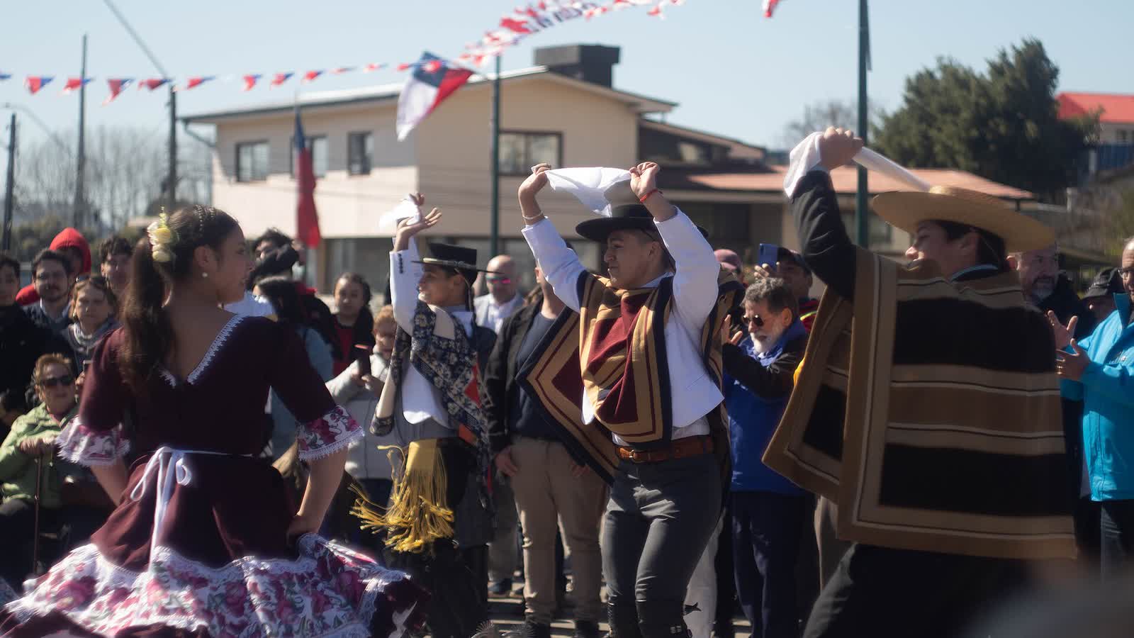 Parejas bailando cueca al aire libre con vestimenta tradicional, pañuelos en alto y banderas chilenas de fondo