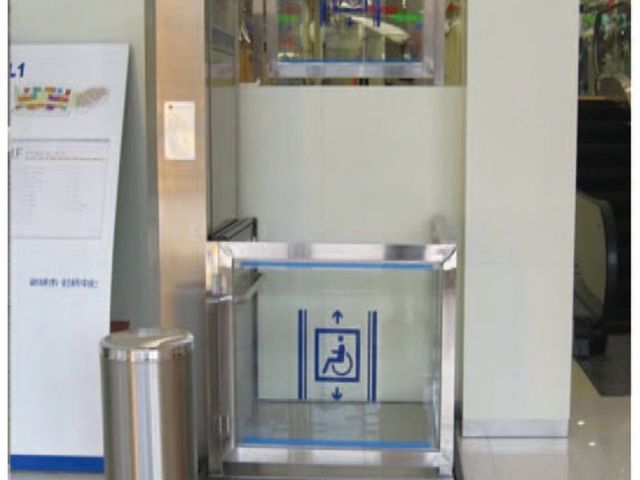 Die Hebebühne in einem Einkaufszentrum: Plattform mit Glaswänden und Rollstuhlzeichen, oben die Haltestelle im Obergeschoss.